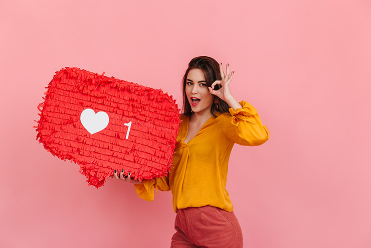 Positive girl in bright blouse and trousers holds like from social media on pink background and shows OK sign