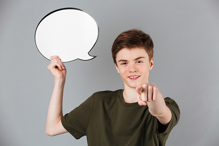 Portrait of a smiling male teenager wearing t-shirt holding blank speech bubble and pointing finger at camera isolated over gray background