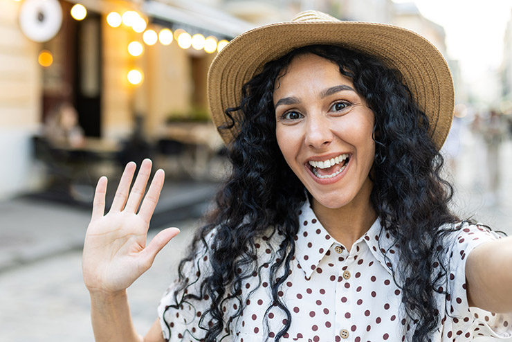 Young beautiful hispanic woman smiling and looking into phone camera, traveler uses smartphone app for remote communication video call, walks in evening city, on tourist tour trip