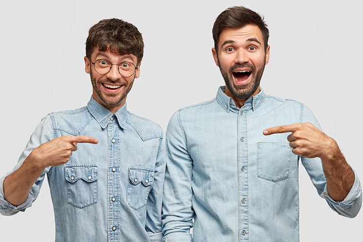 Waist up portrait of two cheerful men indicate with fore fingers at each other, happy to buy new denim jackets, pose against white studio wall, involved in promotion. People, happiness, advertisement