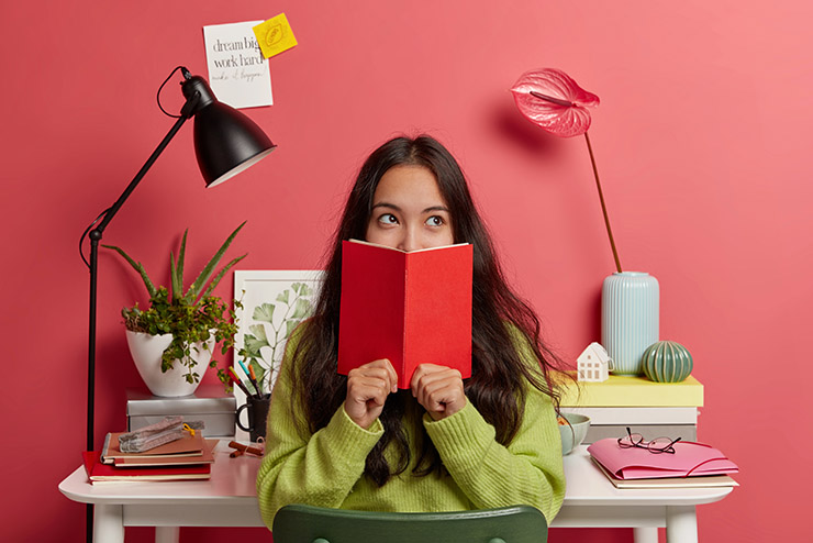 Beautiful brunette pensive mixed race female student learns information from textbook, covers half of face with red diary, poses in coworking space against desktop with necessary things for studying