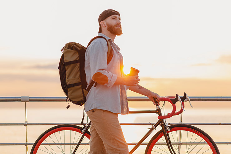 handsome hipster style bearded man with backpack wearing denim shirt and cap with bicycle in morning sunrise by the sea drinking coffee, healthy active lifestyle traveler backpacker